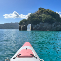 Kayaking in New Zealand Photo of the front of a sea kayak on cerulean blue water looking at a rock cropping with a hole in it.