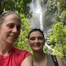 Jen and Amy at a waterfall in NZ The photo shows the head and shoulders of two women in front of a waterfall in the woods of New Zealand. The woman on the left is blond with sunglasses on her head and a red, short-sleeved t-shirt. The woman on the left has dark hair with sunglasses on her head and a brown tanktop and backpack.